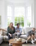 Family sitting on a light-colored sofa in a bright living room with two dogs, plants, and modern art on the walls.