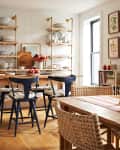 Dining room with wooden table, woven chairs, open shelves, blue stools, and framed botanical prints on the wall.