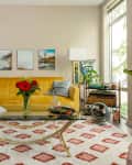 Living room with yellow sofa, glass coffee table, red flowers, and wall art, featuring large windows and a patterned rug.
