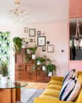 Living room with yellow sofa, glass coffee table, plants, and wall art on peach walls.