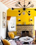 Bright living room with yellow walls, patterned fireplace, modern chandelier, and colorful cushions on a sectional sofa.