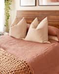 Cozy bedroom featuring a wooden headboard, pink bedding, large pillows, a braided blanket, and a small side table with decor.