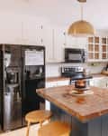 Rustic kitchen with brick island, black fridge, white cabinets, brass pendant light, and red patterned rug.