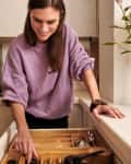 Woman organizing kitchen drawer with utensils, cat sitting on counter, vase of flowers nearby.