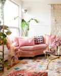 Cozy living room with pink sectional sofa, colorful pillows, macramé wall hanging, plants, and a glass coffee table.