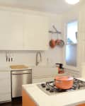 Modern kitchen with white cabinets, orange pot on stove, hanging pans, and decorative vases on counter.