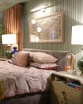 Cozy bedroom with patterned bedding, green lamps, abstract art, and warm lighting. Orange curtains frame the windows.