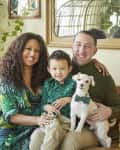 Family sitting on a brown sofa with a small white dog, surrounded by green plants and colorful cushions.