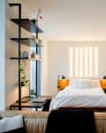 Modern bedroom with a white bed, wooden nightstands, vertical light fixture, and black shelving with plants.