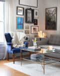 Cozy living room with a gray sofa, blue armchair, gallery wall, glass coffee table, and candles on a side table.
