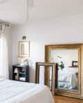 Large gold-framed mirrors leaning against the wall in a simple bedroom with white bedding