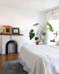 Cozy bedroom with white bedding, potted plant, vintage fireplace, and wooden dresser.