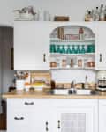 White kitchen with wooden countertop, coffee maker, fruit bowl, and open shelves displaying glasses and bottles.