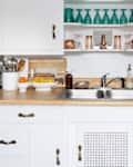 Bright kitchen countertop with a sink, coffee maker, fruits in bowls, and glassware on shelves.
