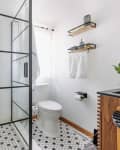 Modern bathroom with glass shower, hexagonal tile floor, wooden vanity, potted plant, and wall shelves with towels.