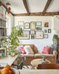Cozy living room with a tan sofa, colorful pillows, plants, bookshelves, and a gallery wall under exposed wooden beams.