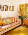 Retro living room with mustard geometric wallpaper, pink sofa, colorful cushions, wooden sideboard, and vibrant plants.