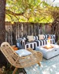 Outdoor patio with striped sofa, wicker chairs, fire pit, and umbrella on a patterned rug under a tree canopy.
