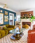 Living room with green sofa, pink velvet chair, glass coffee table, brick fireplace, and floral decor on a blue accent wall.