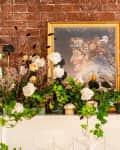 Floral arrangement with white roses and greenery on a mantel, framed painting on a brick wall backdrop.