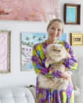 Woman in colorful dress holding a fluffy cat, standing in front of a gallery wall with various framed artworks.