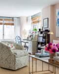 Cozy living room with a patterned armchair, bookshelves, floral decor, and a desk by the window with natural light.