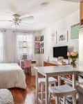Cozy studio apartment with a bed, sofa, white shelves, pink chair, and wall art, featuring a TV and a small dining area.