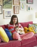 Woman sitting on a red sofa with colorful pillows, holding a cat, surrounded by framed art on the wall.