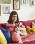 Woman sitting on a red sofa with colorful pillows, holding a cat, surrounded by framed art on the wall.