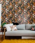 Gray sofa with colorful floral pillows against a vibrant floral wallpaper, wooden floor, and yellow rug.