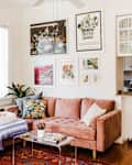 Cozy living room with a pink sofa, colorful pillows, wall art, and a small kitchen nook with open shelving.