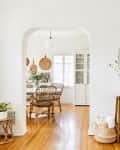 Entryway with a round mirror, console table, and vase of flowers, leading to a dining room with wicker chairs and wooden floor.