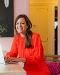 Woman in red dress using a laptop at a table with a small dog, flowers, and abstract art on a pink wall.