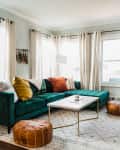 Green velvet sectional sofa with colorful pillows, white coffee table, poufs, and plants in a bright living room.