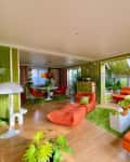 Retro living room with orange and green decor, patterned wallpaper, and modern furniture, featuring plants and a dining area.