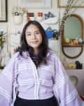 Woman in a room with eclectic art on walls, wearing a purple striped jacket, standing in front of a shelf with decor items.