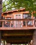 Treehouse with wooden deck, surrounded by green leaves, featuring a small table and chairs.