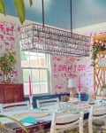 Bright dining room with pink patterned wallpaper, a wooden table, white chairs, and a rattan shelf displaying decor and plants.