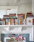 Books stacked on a white mantel with framed photos, plants, and a drawing of a cocktail.