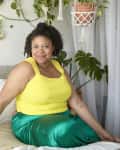 Woman in a yellow top and green skirt sitting on a bed in a plant-filled room with a cozy, bohemian decor.