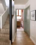 Hallway with light wood flooring, leading to a bathroom with a wooden vanity and a window. Beige carpeted stairs on the left.