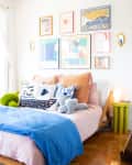 Cozy bedroom with colorful pillows, blue duvet, wall art, and a small green side table with a lamp.