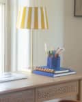 Desk with a laptop, potted plant, striped lamp, and stationery holder by a window.