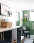Bathroom with green walls, black chair, hanging plant, wicker basket, and framed art on shelf.