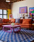 Loft living room with orange sectional sofa, colorful abstract art, purple coffee table, and blue checkered rug.