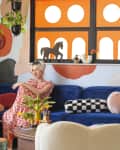 Colorful living room with blue velvet sofa, patterned pillows, abstract wall art, and a woman in a red dress sitting with a cat.