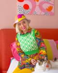 Woman in colorful outfit sitting on bed with cat, surrounded by vibrant decor and abstract wall art.