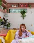 Woman in a cozy living room with yellow sofa, pink walls, and lush green plants.