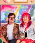 Couple sitting on a sofa with a cat, surrounded by colorful art and decor, including vibrant pillows and flowers.