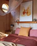 Cozy bedroom with wood paneling, round window, pink bedding, and a shelf with candles and art above the bed.